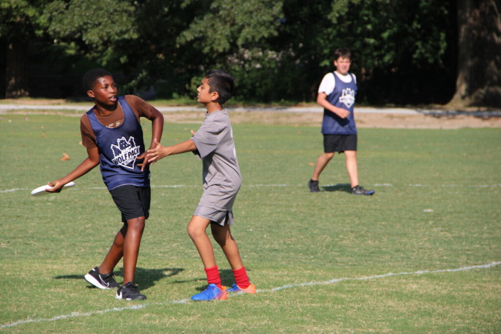 Two middle school students compete during an ultimate frisbee game on a sunny grass field. The student on the left, wearing a navy Wolf Pack Ultimate pinnie, holds a frisbee while being defended by a student in a gray shirt and bright red socks. A third student in a Wolf Pack pinnie is visible in the background. Lush green trees line the edge of the field behind them.