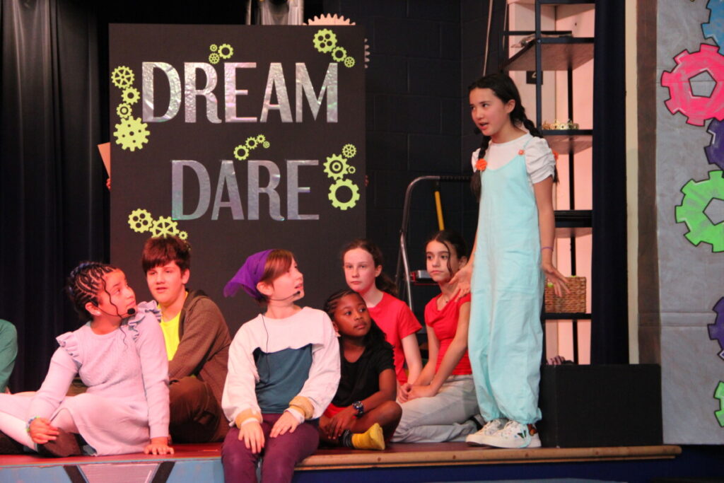 A group of middle school students perform on a theatrical stage. One student stands center-right in a light blue jumpsuit, speaking or singing while her castmates sit and look up at her attentively. Behind them, a large black backdrop displays the words "Dream Dare" in silver metallic letters surrounded by yellow gear illustrations. Colorful oversized gear decorations are visible on the right side of the stage.