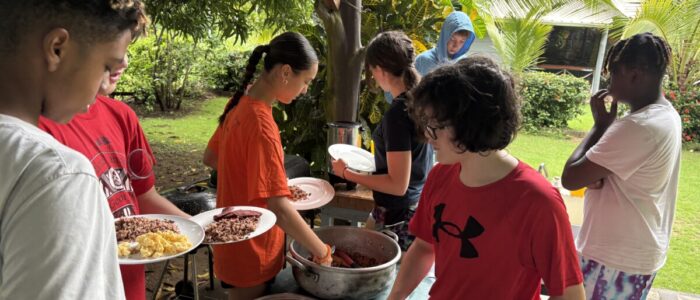 Costa_Rica_2026_Food A group of TCS middle school students serve themselves a traditional Costa Rican breakfast of rice, beans, scrambled eggs, and plantains from large pots set up on an outdoor table, surrounded by tropical greenery.