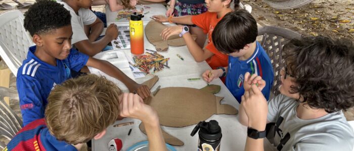 Costa_Rica_2026_Leadership TCS middle school students sit around an outdoor table concentrating on an art project, using crayons to decorate large cardboard cutouts shaped like sea turtles. A hammock and lush green grass are visible in the background.
