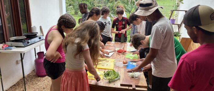Costa_Rica_2026_Food_Prep A large group of TCS middle school students crowd around a wooden table on an open-air patio in Costa Rica, chopping and preparing fresh vegetables including peppers, onions, and greens during a cooking activity. A portable gas stove is visible to the left, and dense tropical forest fills the background.