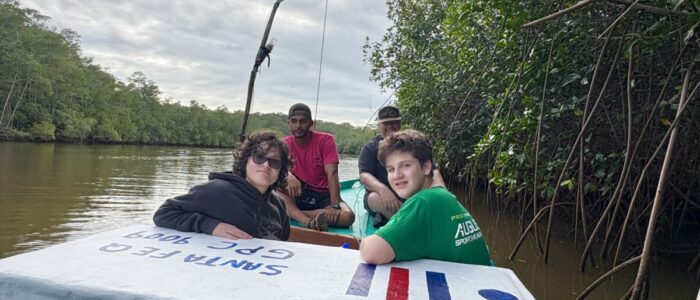 Costa_Rica_2026_Mangrove Two TCS students lean against the bow of a small fishing boat navigating a narrow, murky river flanked by dense mangrove trees in Costa Rica. Two adult guides sit behind them under a cloudy sky.