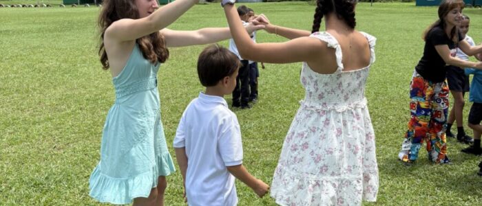 Costa_Rica_2026_School Two TCS middle school students in sundresses hold hands with a young Costa Rican boy in school uniform on a wide green field, playing a traditional game or folk dance together. Other students and children are visible playing in the background, with forested hills rising behind them.