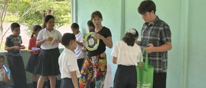 Costa_Rica_2026_Gifts A TCS middle school student and a chaperone distribute gifts including toys and a frisbee to Costa Rican elementary students in school uniforms inside a covered outdoor school corridor. Trees are visible through the open walls behind them.