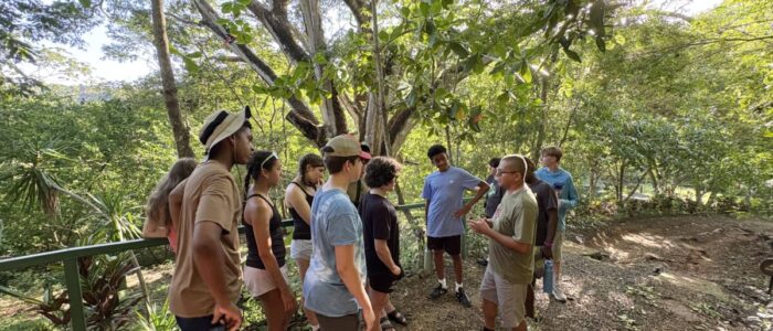 Costa_Rica_2026_Forest A group of TCS middle school students gathers in a shaded forest clearing in Costa Rica, listening attentively as a local guide speaks to them beneath a large canopy of tropical trees with sunlight filtering through the leaves.
