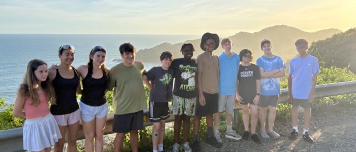 Costa_Rica_2026_Group TCS middle school students stand together along a roadside guardrail with a breathtaking golden sunset, sparkling ocean, and rolling green coastal mountains stretching behind them in Costa Rica.