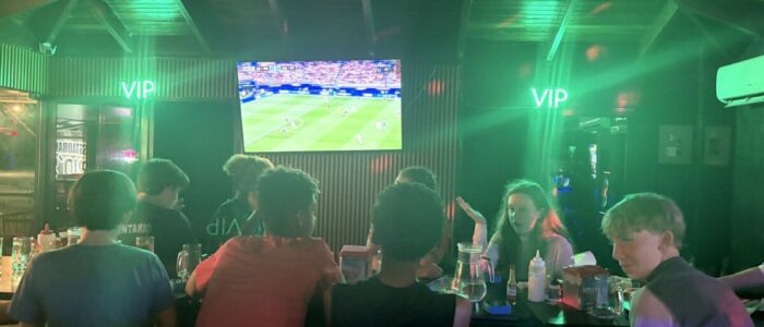 Costa_Rica_2026_TV TCS middle school students sit together at tables inside a dimly lit Costa Rican restaurant with green neon lighting, watching a soccer match broadcast on a large screen mounted on the wall.
