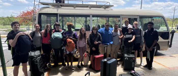 Costa_Rica_2026_Bus TCS middle school students, chaperones, and a local guide stand together in front of a passenger van loaded with luggage on its roof rack, gathered outside in a sunny Costa Rican parking area at the start or end of their trip.