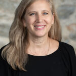 Professional headshot of Alison Schers, Director of Enrollment at The Children's School. She is smiling warmly, wearing a black top and gold jewelry, with long blonde-gray hair, photographed against a soft stone background.