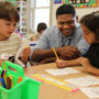 A smiling teacher in a light blue shirt leans in to support two third grade students working on a drawing activity at a shared classroom table. The student on the left examines a piece of paper while the student on the right, wearing red headphones, works on a face outline drawing with a pencil. A green caddy filled with colorful pencils and markers sits in the foreground, with additional students and a bright, well-supplied classroom visible in the background.
