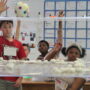A group of middle school students raise their hands enthusiastically in a busy, print-rich classroom. In the foreground, a clear acrylic shelf holds several small white structures made from cotton balls and other World Peace Game materials. A classroom calendar, whiteboard with notes, and student work cover the walls in the background.