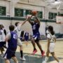 A player wearing a blue #31 jersey leaps to shoot over a defender in a white jersey (#25) during an indoor middle school basketball game. Other players in blue and white uniforms are visible on the court, with championship banners and bleachers in the background.