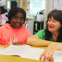 A student in a pink hoodie smiles at the camera while sitting at a classroom table with an open book in front of her. A teacher in a green shirt sits beside her, smiling warmly as she looks down at the book. Other students are visible in the background of the bright, busy classroom.