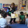 An educator crouches down to show an open picture book to a small group of young students seated on a colorful rug in a bright, print-rich kindergarten classroom. The walls behind her are covered with learning materials including a November calendar, vowel charts, number displays, and colorful classroom schedules. The children look attentively at the book being shared with them.