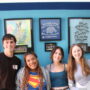 Four smiling teenagers stand together in front of a bright teal wall displaying a gallery of framed graduating class posters from The Children's School. The students are casually dressed and posed closely together, smiling at the camera. The framed artwork behind them includes class names, colorful designs, and TCS Wolf Pack imagery.