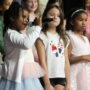A young student in a white cardigan and pastel tutu skirt holds a microphone and sings during a school performance. She stands at the front of a group of elementary-aged students on a stage, several of whom are dressed in festive outfits including a sparkly pink dress and a sequined top. Additional students are visible standing in rows behind them.