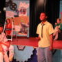 A middle school student in a yellow button-down shirt and jeans performs center stage, speaking or singing while a large cast of fellow students watch from tiered risers behind him. The elaborately decorated stage features a giant gear prop, a colorful collage backdrop with movie posters and illustrations, and a blue panel on the right displaying a Walt Disney quote about dreams and courage. Colorful gear decorations line the left side of the stage.